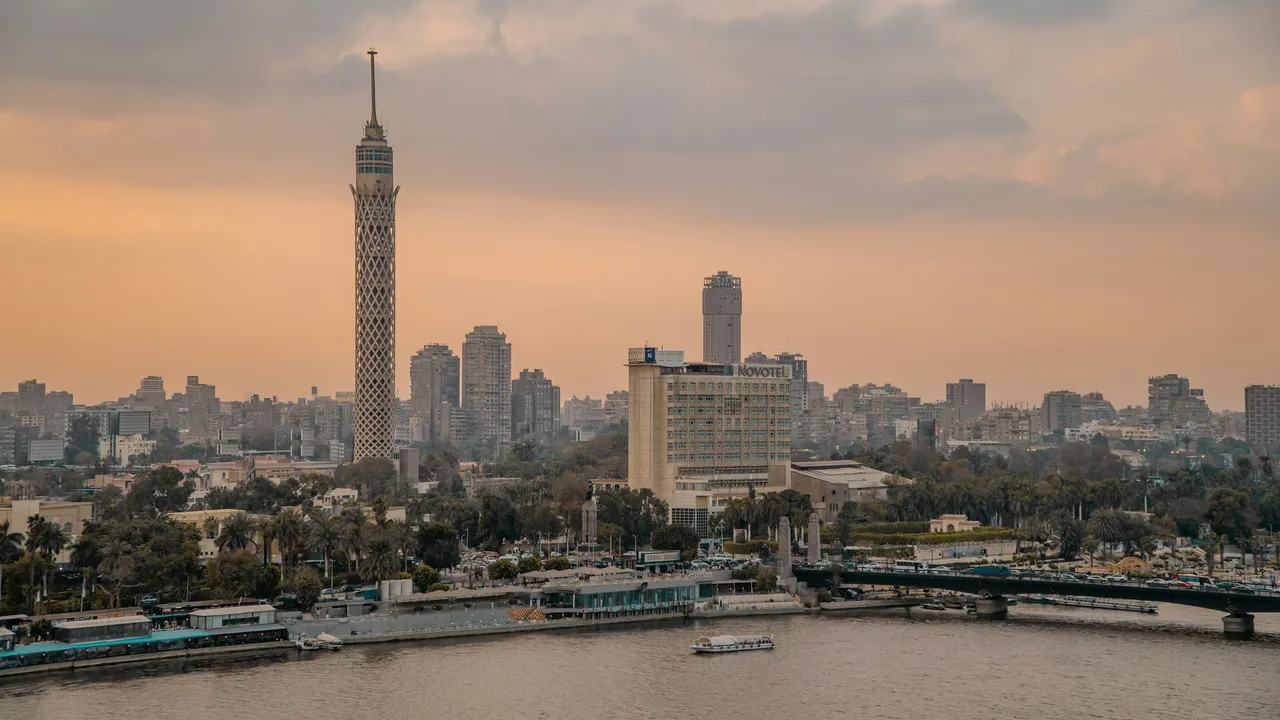 A wide view of the Cairo skyline in Egypt along the Nile River during sunset. The prominent lattice-work Cairo Tower stands tall on the left, with the Novotel hotel and other urban buildings lining the riverbank under a warm, hazy sky.