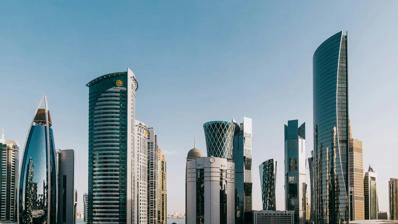 A wide, low-angle shot showcases the modern, gleaming skyline of Doha, Qatar, under a clear blue sky. The city's architectural diversity is on display, featuring a variety of glass-faced skyscrapers, including a prominent curved tower with a diamond-patterned exterior and others with sharp, geometric finishes.