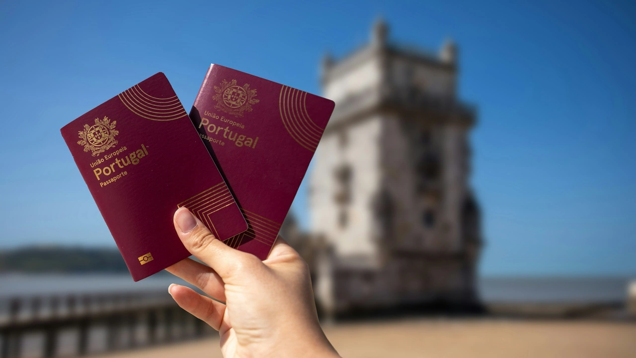 A close-up shot shows a hand holding two burgundy Portuguese passports in the foreground. The background is a sunny, blurred view of the historic Belém Tower in Lisbon, situated along the waterfront under a clear blue sky.