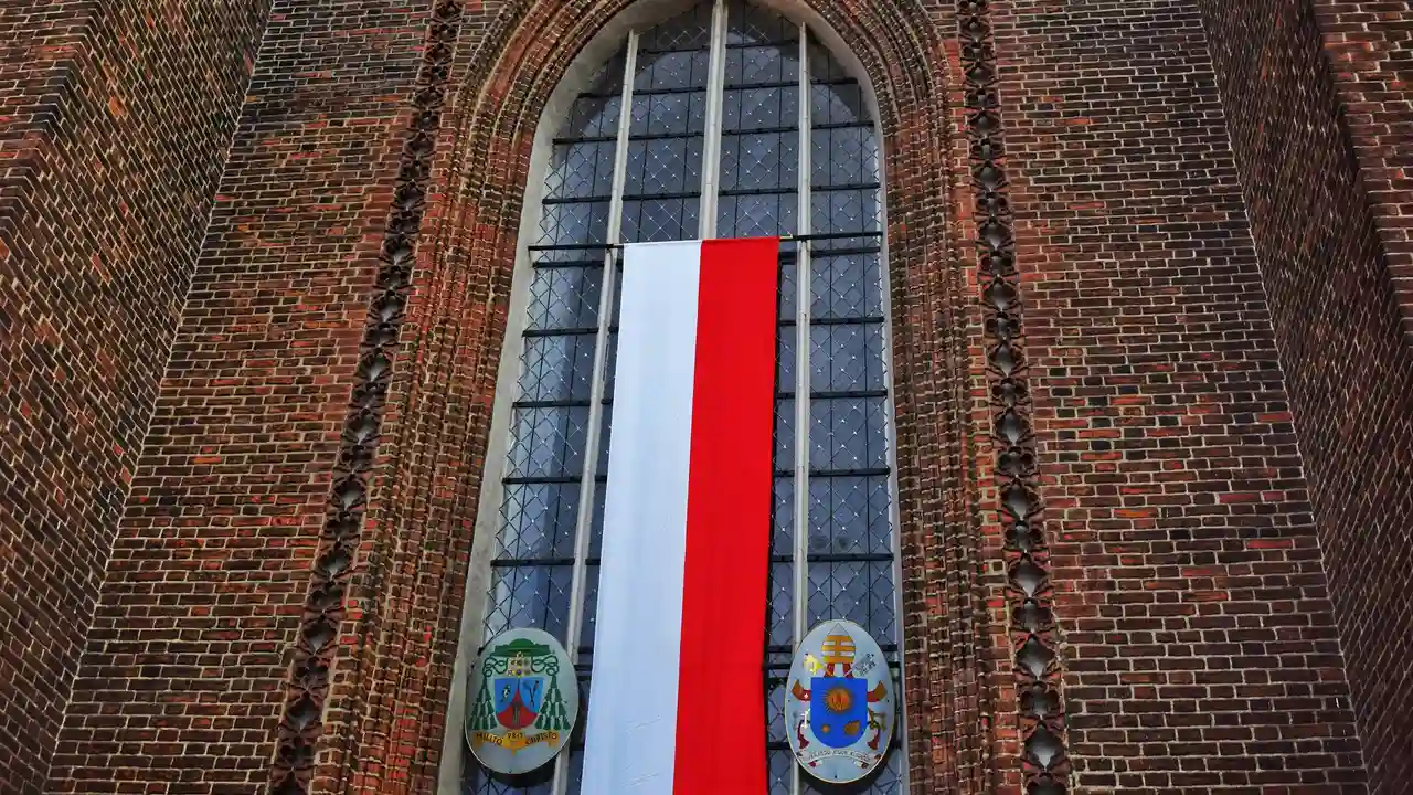 A vertical Polish flag hangs in front of a tall, arched stained-glass window of a historic brick building. At the base of the window, two decorative circular emblems are displayed on either side of the flag's red and white stripes.
