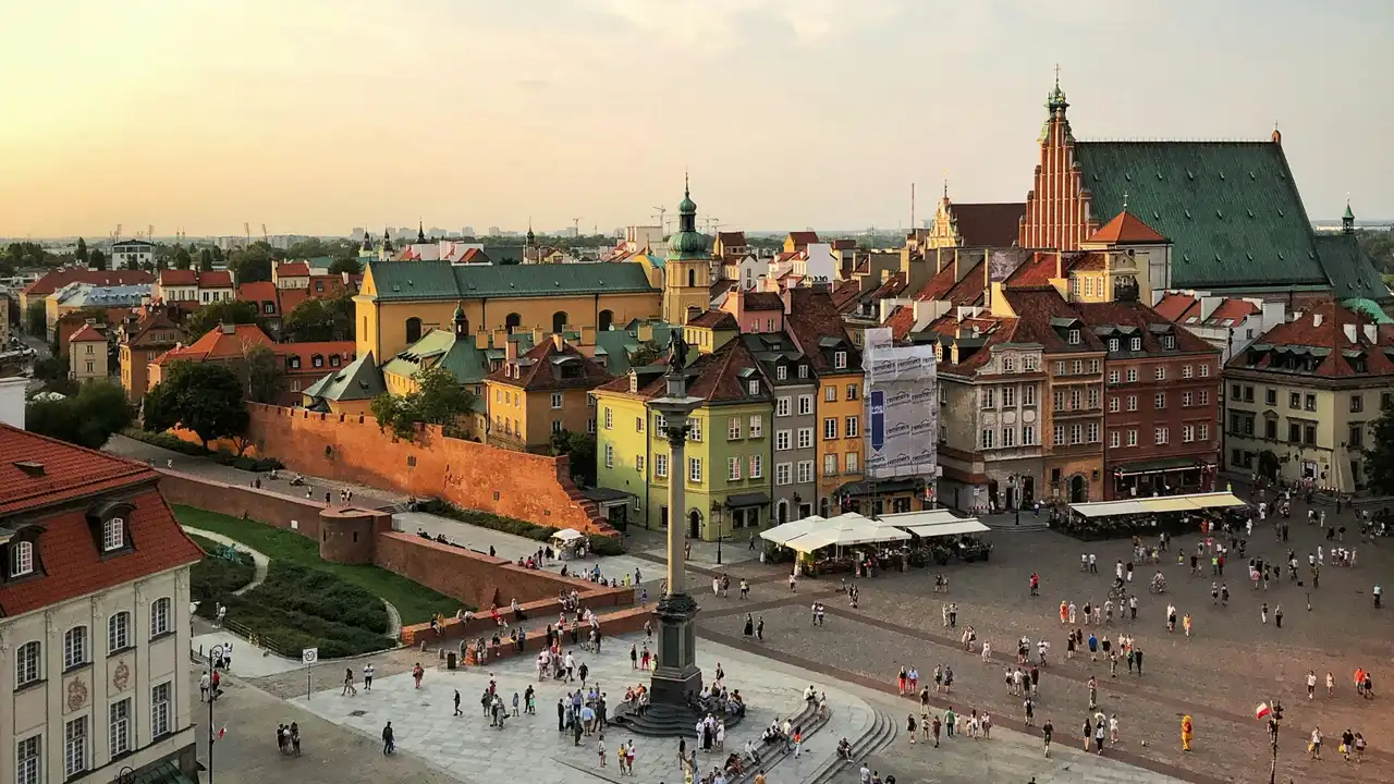 An elevated, high-angle view of Warsaw's Castle Square in Poland during the late afternoon. The scene features a large, paved plaza filled with pedestrians and a tall central monument, flanked by a long brick fortification wall and a row of colorful historic buildings with varied rooflines and a prominent church tower in the background.