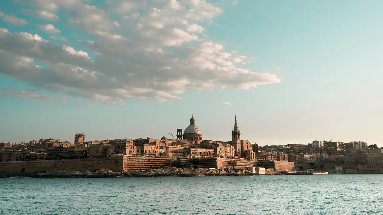 A wide-angle, scenic view of the Valletta skyline in Malta across the Mediterranean Sea. The historic city features golden limestone buildings, ancient fortifications, and the prominent dome and spire of the Basilica of Our Lady of Mount Carmel. The sky is a clear light blue with soft, scattered clouds, and the turquoise water in the foreground is calm.