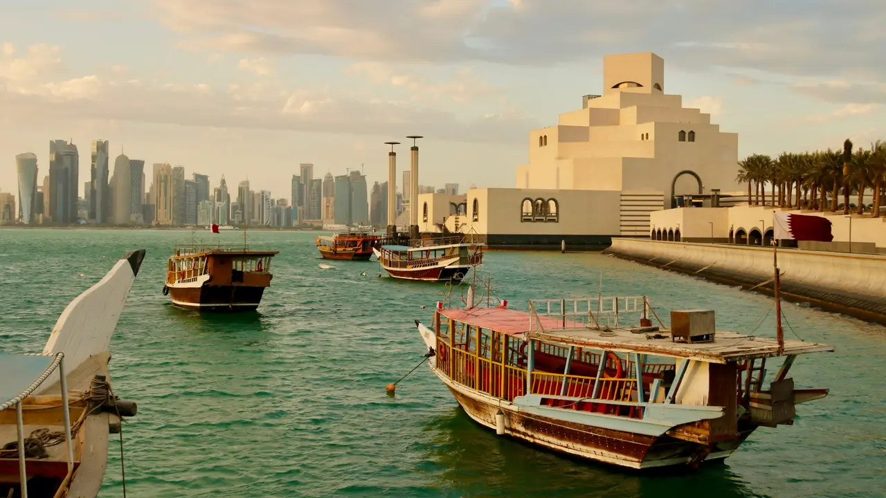 A high-angle scenic view shows several traditional wooden dhow boats floating on calm, turquoise water in Doha, Qatar. In the background, the distinctive white, geometric architecture of the Museum of Islamic Art stands on the waterfront, with the modern city skyline visible in the distance under a soft, late-afternoon sky.