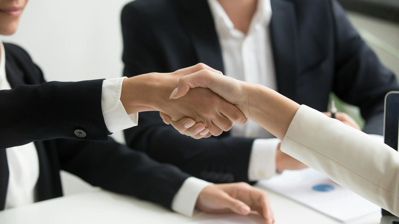 A close-up, high-angle shot of two people's hands shaking over a dark wooden office table. One person is wearing a light blue dress shirt, and the other is wearing a white dress shirt with a dark suit jacket. The handshake is centered in the frame, symbolizing a professional agreement or successful meeting.