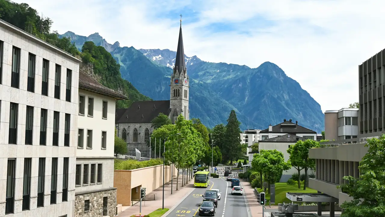  An eye-level view of a street in Vaduz, Liechtenstein, featuring the Cathedral of St. Florin with its tall, dark spire. Modern buildings and green trees line the road where a yellow bus and several cars are driving, all set against a backdrop of steep, lush green mountains under a blue sky.