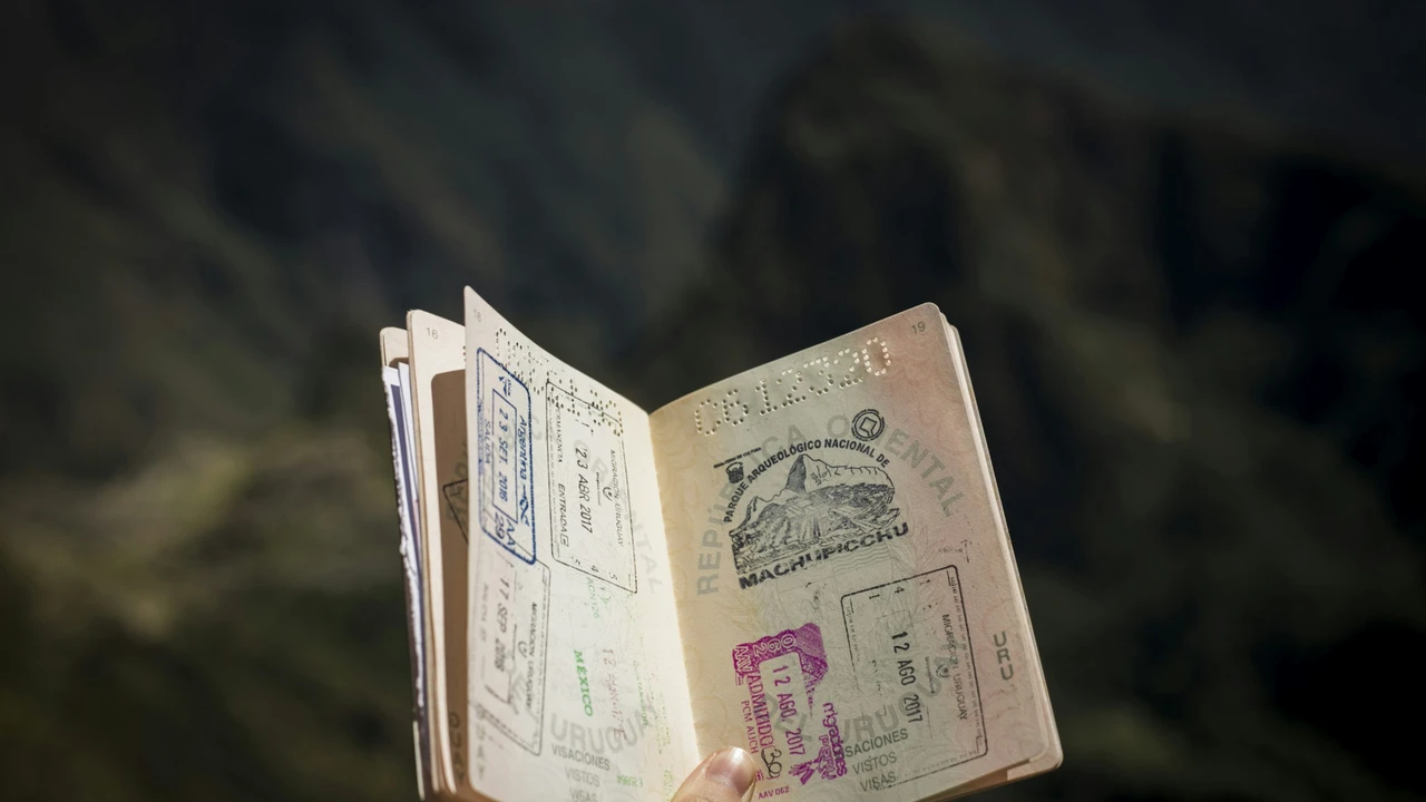 An open passport is held up against a blurry, dark green and brown mountain background. The passport pages are filled with various colorful travel stamps, including a large, prominent black stamp from the Machu Picchu archaeological site in Peru and others from Uruguay, Argentina, and Mexico.