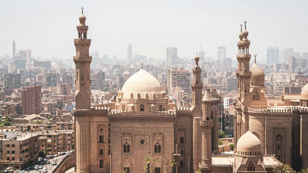 A high-angle view of the historic Mosque-Madrasa of Sultan Hassan in Cairo, Egypt. The image showcases its massive stone walls, intricate domes, and tall minarets against a backdrop of a densely populated city under a bright sky.