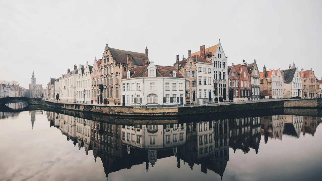 A wide shot of historic Flemish stepped-gable houses lining a canal in Bruges, Belgium, perfectly reflected in the still, glassy water under an overcast sky. A stone bridge and a distant church spire are visible on the left side of the scenic waterway.