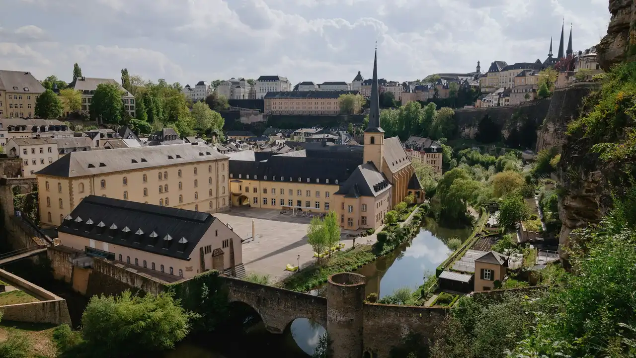 A high-angle view of the Grund district in Luxembourg City, featuring historic stone buildings with slate roofs, a prominent church spire, and an old stone bridge over a narrow river. Lush greenery and rocky cliffs surround the valley.