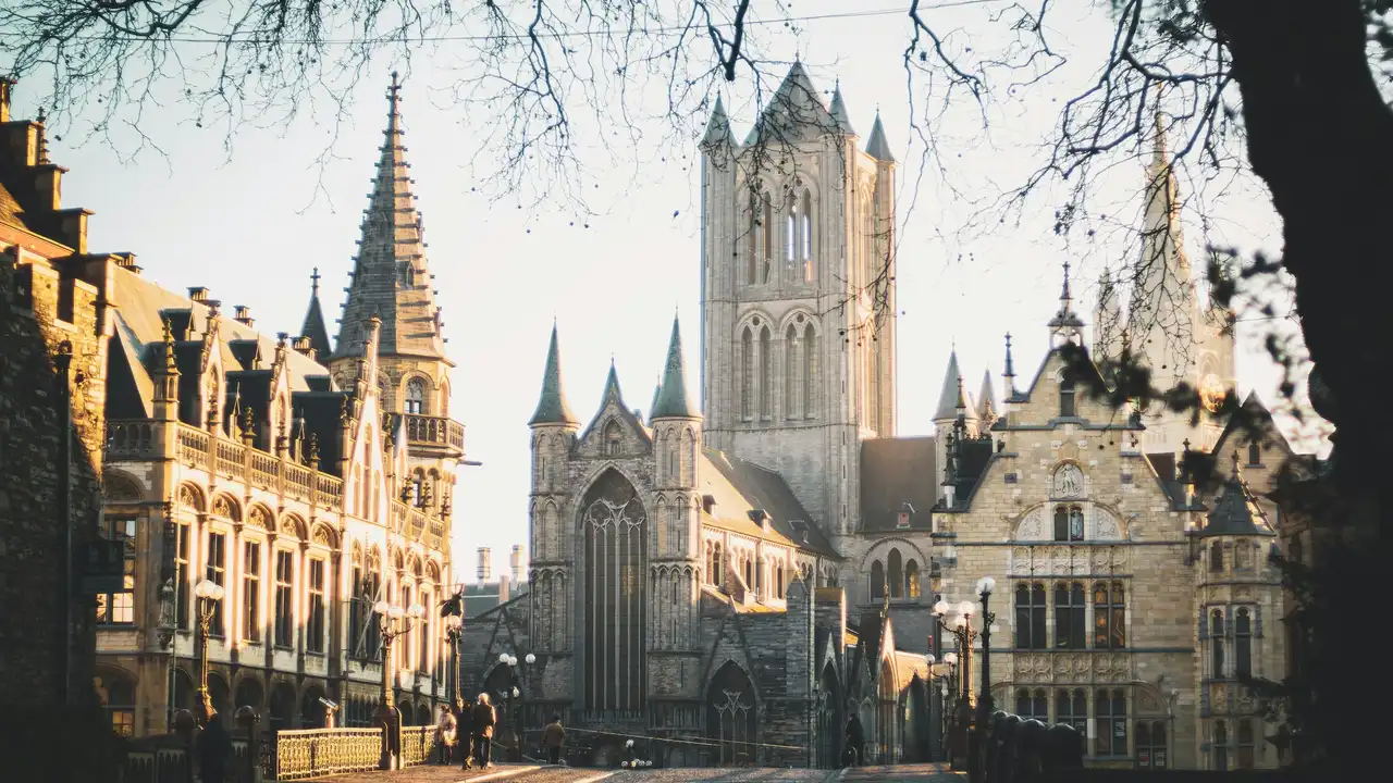 A sunlit view of a historic cobblestone street in Ghent, Belgium, featuring the gothic architecture of St. Nicholas' Church and the Old Post Office. Bare tree branches frame the top of the image as pedestrians walk near the bridge in the soft, golden light.