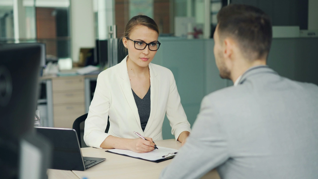 Professional job interview scene with a female recruiter wearing glasses and a white blazer reviewing notes while interviewing a male candidate across a desk in a modern office setting.