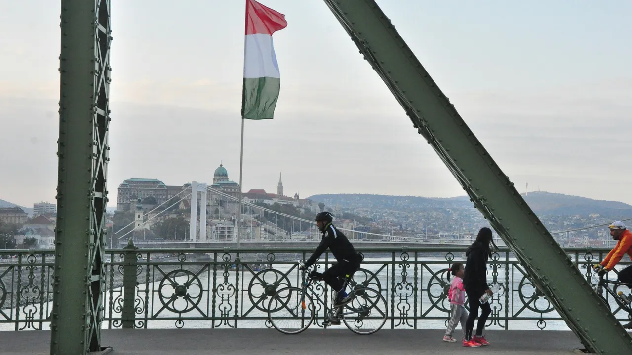 Liberty Bridge in Budapest, Hungary, with the Hungarian national flag flying above, showing cyclists and pedestrians crossing the bridge along the Danube River, with Budapest cityscape and historic buildings in the background.