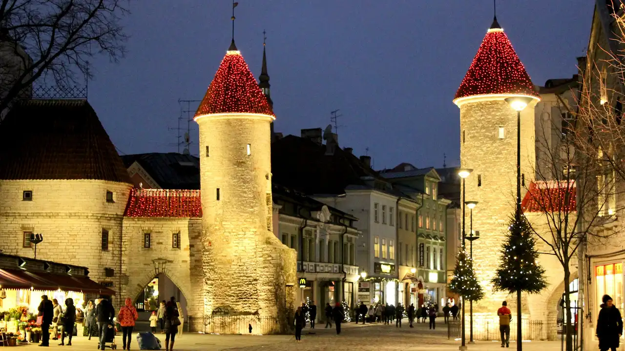 Evening view of Tallinn Old Town in Estonia, featuring medieval stone towers decorated with lights, historic buildings, and people walking through the illuminated street.