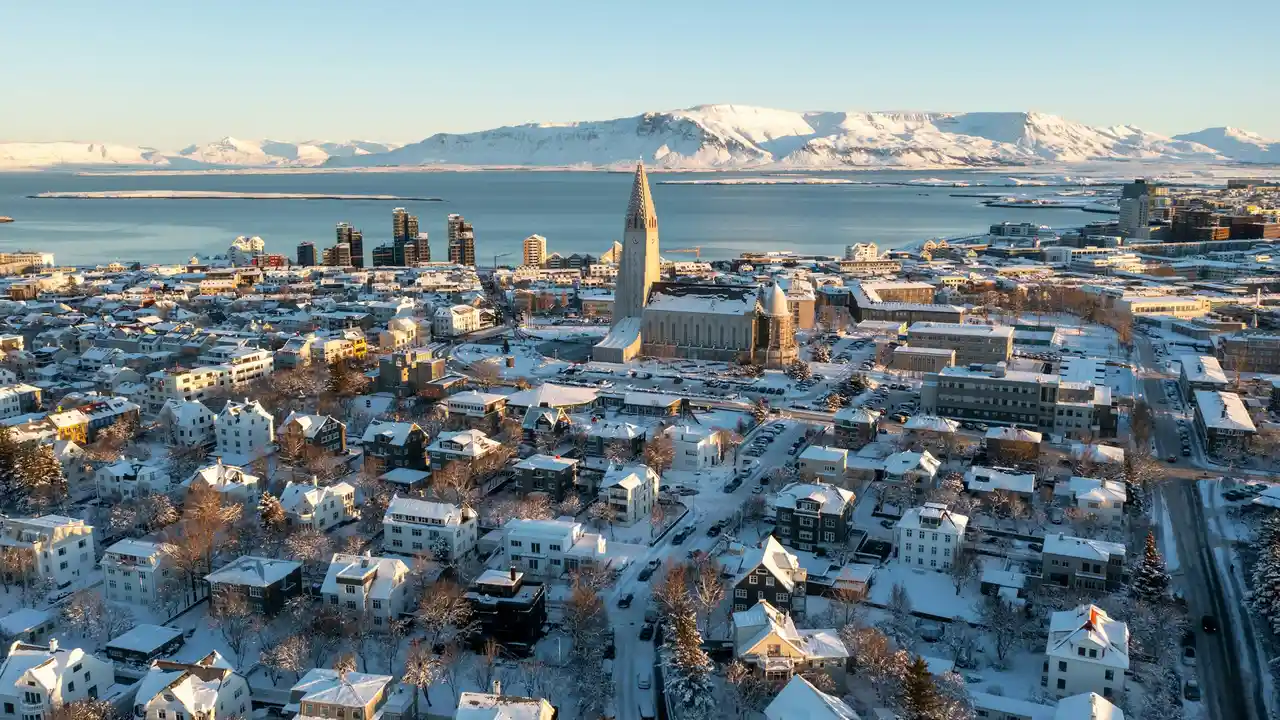 Aerial winter view of Reykjavik, Iceland, showcasing snow-covered houses, Hallgrímskirkja church, coastal scenery, and mountains, highlighting why people want to visit Iceland for its natural beauty and unique cityscape.
