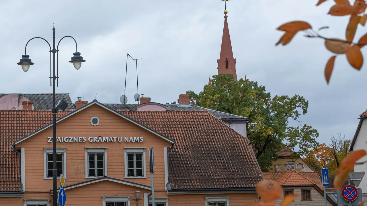 A medium shot of an peach-colored wooden building with a red-tiled roof in Cēsis, Latvia. The text 