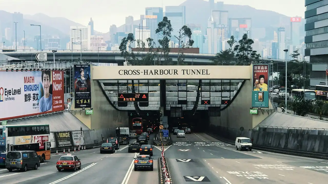 Do Indians Need A Visa To Visit Hong Kong A wide-angle shot of the entrance to the Cross-Harbour Tunnel in Hong Kong, showing multiple lanes of traffic with cars, taxis, and a double-decker bus. Large advertising billboards are visible on either side of the tunnel entrance, and the city's dense skyline of skyscrapers rises against a hazy mountain backdrop in the distance.