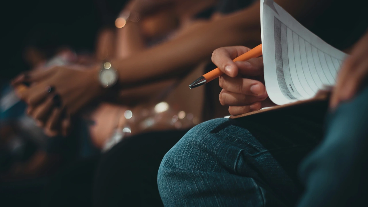 A close-up shot focused on a person's hand holding an orange pen over a clipboard with a document. The person is sitting in a crowded room with other people visible in the blurred background, suggesting a setting like a lecture, conference, or waiting area where notes or applications are being completed