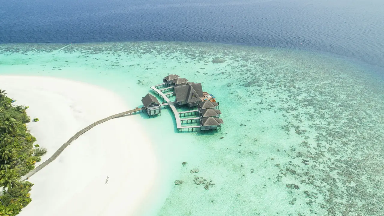 Aerial view of luxury overwater villas connected to a white sandy beach, surrounded by crystal-clear turquoise ocean, depicting a tropical island resort.