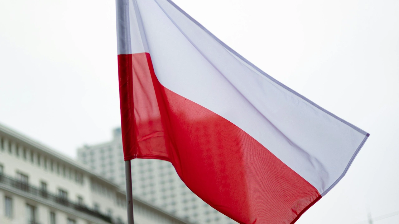 The national flag of Poland, featuring two horizontal stripes of white on top and red on the bottom, waves in the foreground against a blurred background of a white building and a grey sky.