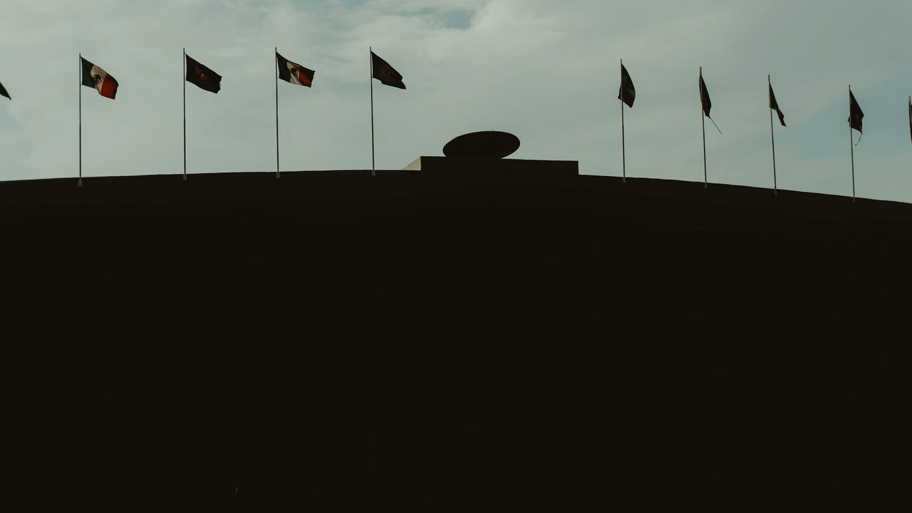 Silhouette of a curved government-style building with multiple national flags flying along the roofline against a cloudy sky.