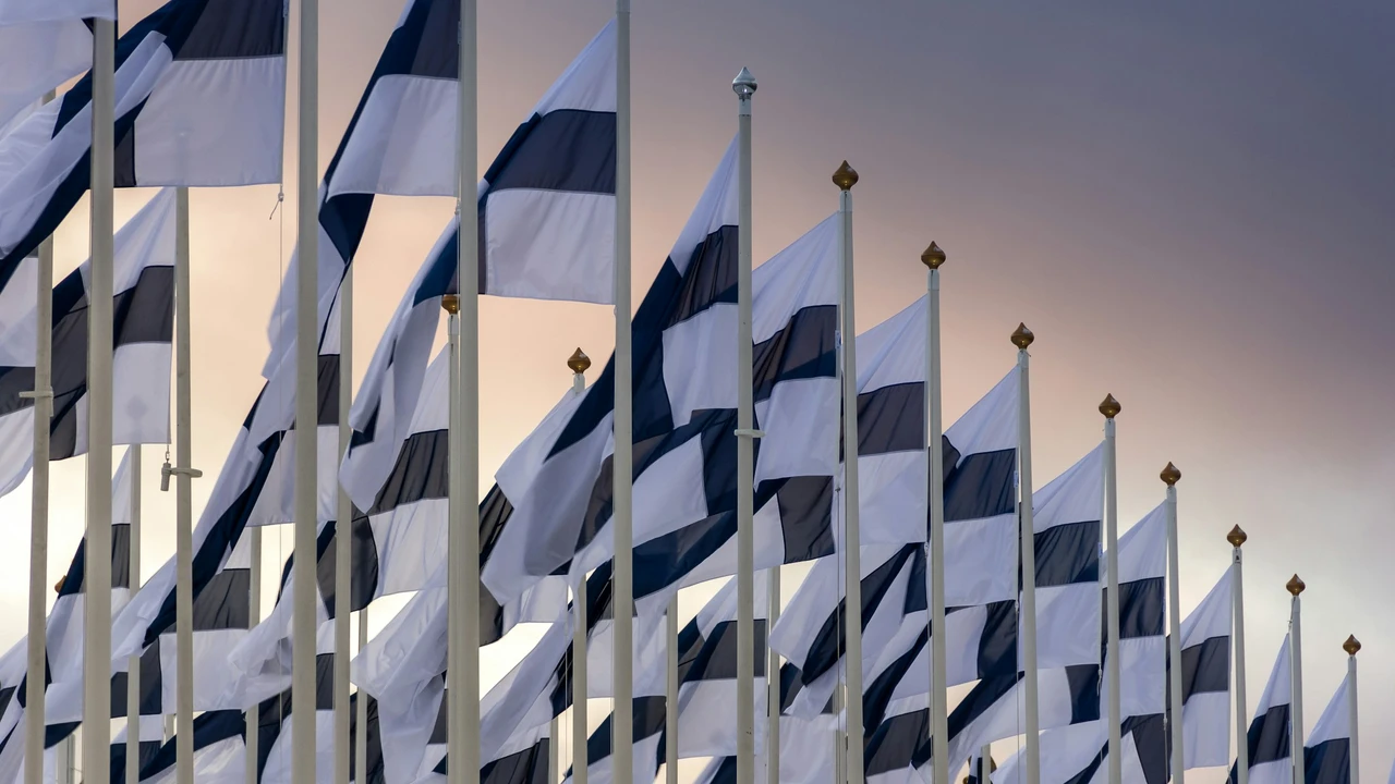 A line of Finnish flags, featuring blue crosses on white backgrounds, are mounted on tall white poles with gold-colored tops. The flags wave in various directions against a soft, gradient sky of muted orange and purple during twilight