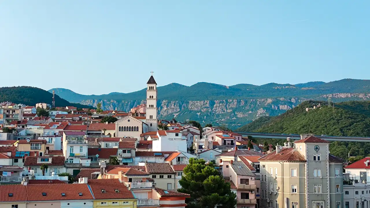 A scenic, wide-angle shot of a coastal Croatian town nestled against a backdrop of rolling green mountains. The foreground is filled with traditional white buildings and houses with red-tiled roofs, while a tall, white church bell tower with a dark spire stands out as a central landmark. A clear blue sky stretches above the mountain ridge in the distance.