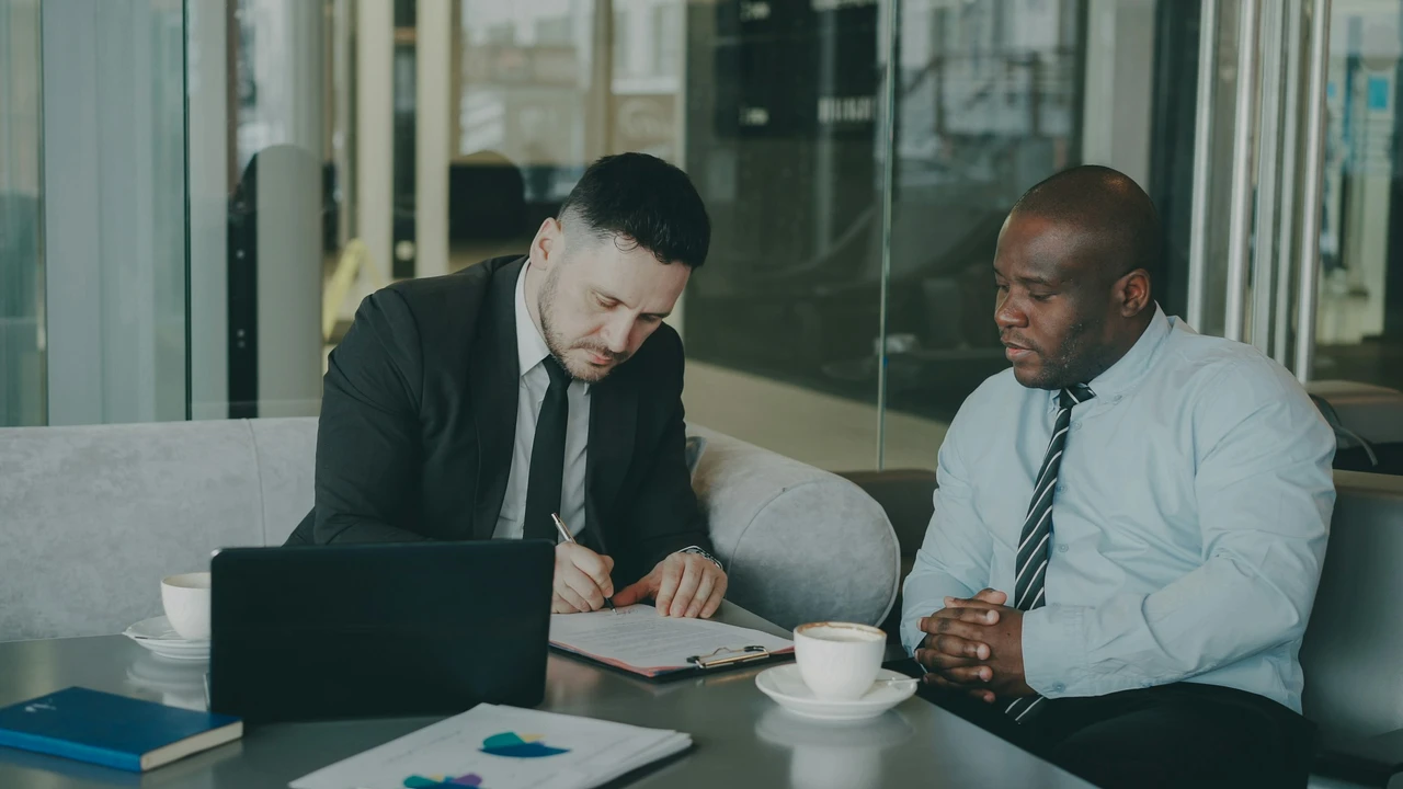 In a modern office or cafe setting, two men in professional attire sit at a grey table. A man on the left, wearing a black suit and tie, is focused on writing on a document held in a clipboard. The man on the right, a Black man in a light blue shirt and striped tie, sits with his hands clasped, watching the process. A laptop, coffee cups, and papers with colorful charts are also on the table
