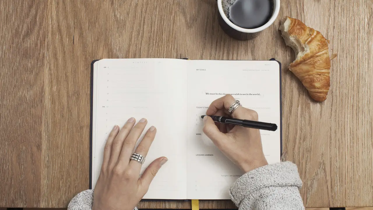 A top-down view of a person’s hands working in an open planner on a wooden desk. One hand wears a silver band, while the other, wearing multiple silver rings, writes with a black felt-tip pen. A black coffee mug and a partially eaten croissant are also on the desk