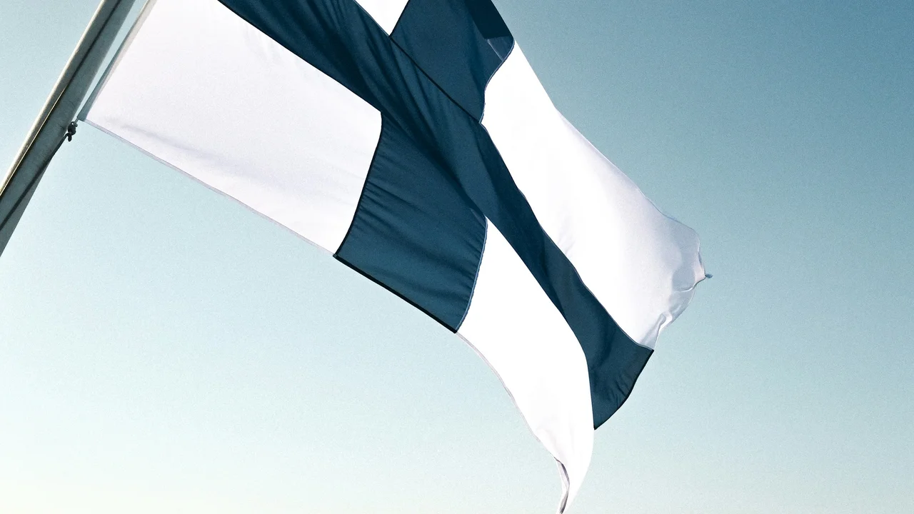 A close-up shot of the Finnish flag, featuring its distinctive blue cross on a white field, waving against a clear, pale blue sky