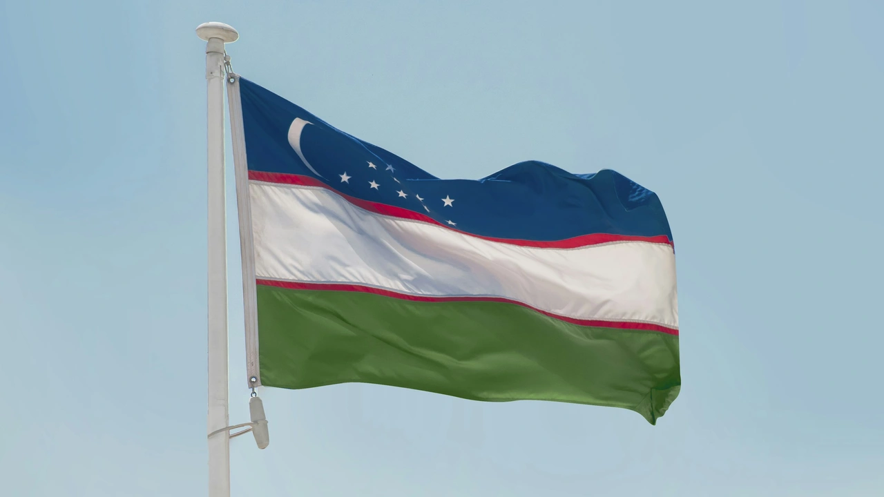 The national flag of Uzbekistan waving on a white flagpole against a clear, pale blue sky. The flag features three horizontal stripes of blue, white, and green, separated by two thin red stripes, with a white crescent moon and twelve small stars in the upper hoist-side corner.
