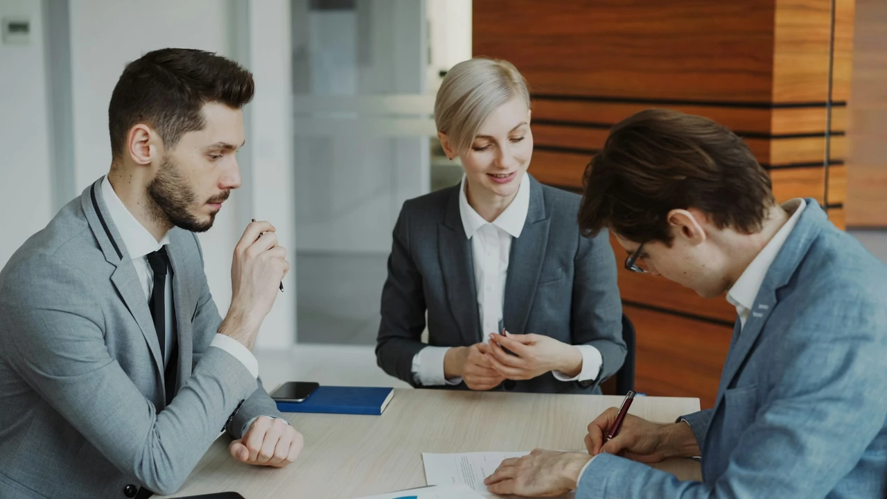 In a professional office environment, three individuals are seated at a light-colored wooden desk. A woman with short blonde hair in a grey suit sits in the center, smiling and engaging with two men. The man on the left, in a grey blazer, listens attentively with a pen in hand, while the man on the right, also in a blue blazer, is focused on signing a document