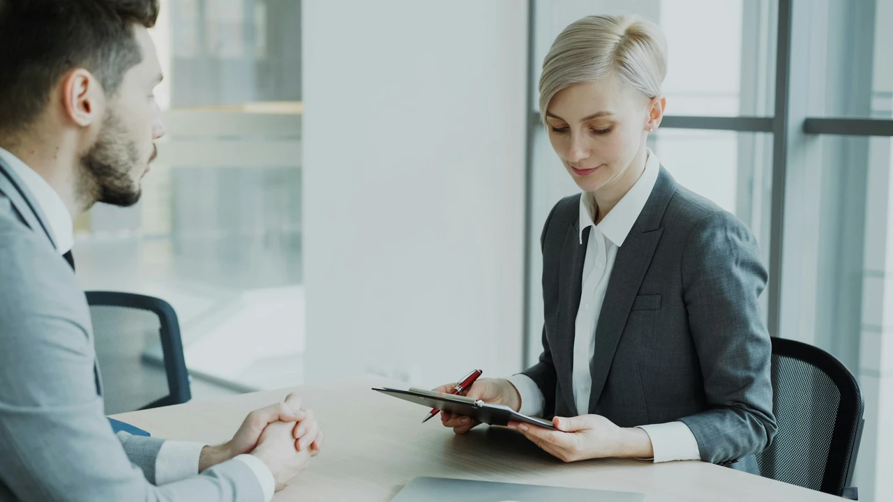 A woman with short blonde hair wearing a grey business suit sits at a desk, looking down at a clipboard while holding a red pen. Across from her, a man in a grey suit sits with his hands clasped on the table, likely participating in an interview or professional consultation.