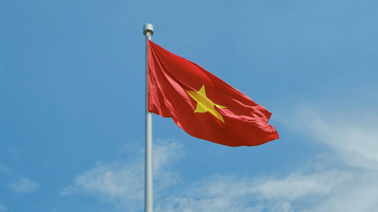 The national flag of Vietnam, featuring a bright red background with a large yellow five-pointed star in the center, waves from a tall silver flagpole against a clear blue sky with light, wispy clouds.