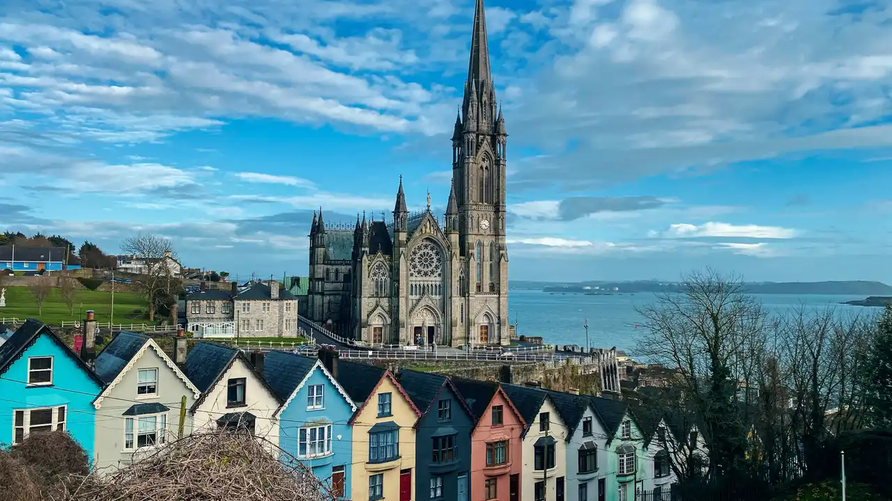 An wide-angle view of the St. Colman's Cathedral in Cobh, Ireland, towering over a row of iconic, multicolored houses known as the