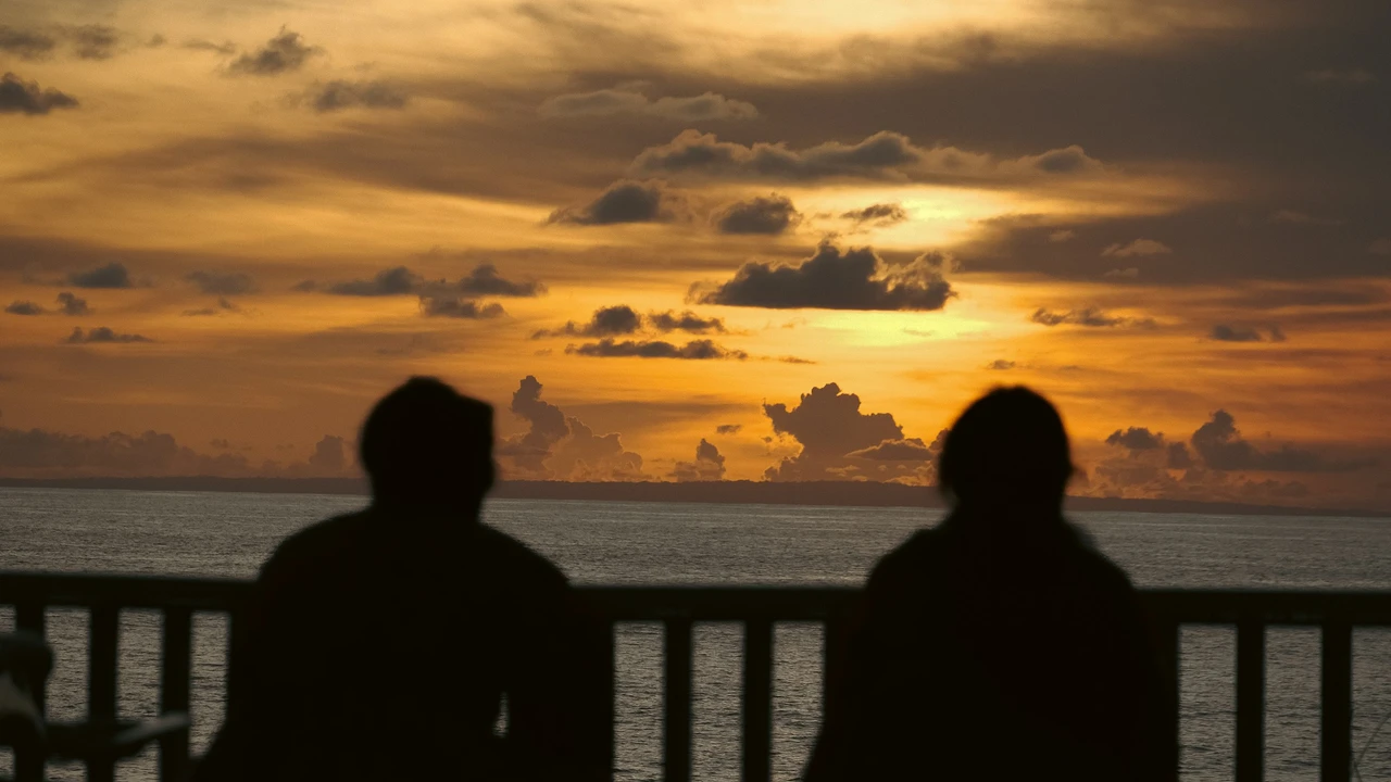 Silhouettes of two people watching dramatic sunset over the ocean from balcony with golden sky and clouds creating romantic evening seascape scene
