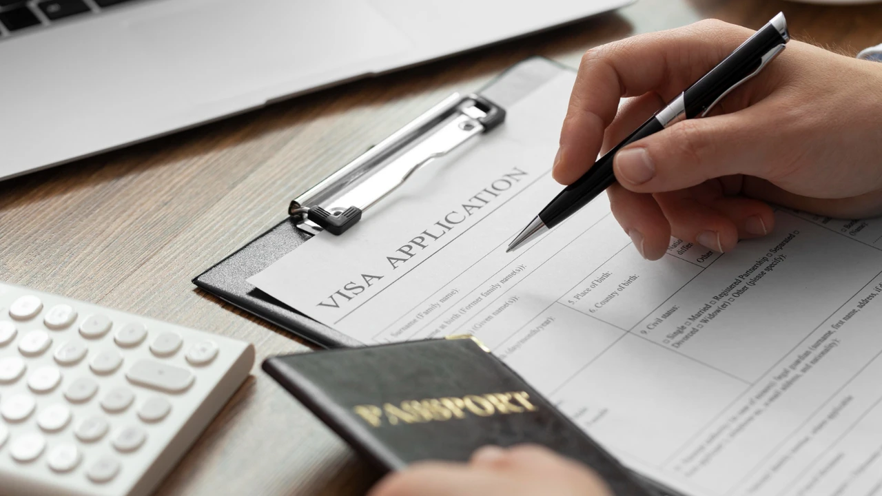 Close-up of a person filling out a visa application form on a clipboard with a pen, passport and calculator on desk, representing travel documentation and immigration process.