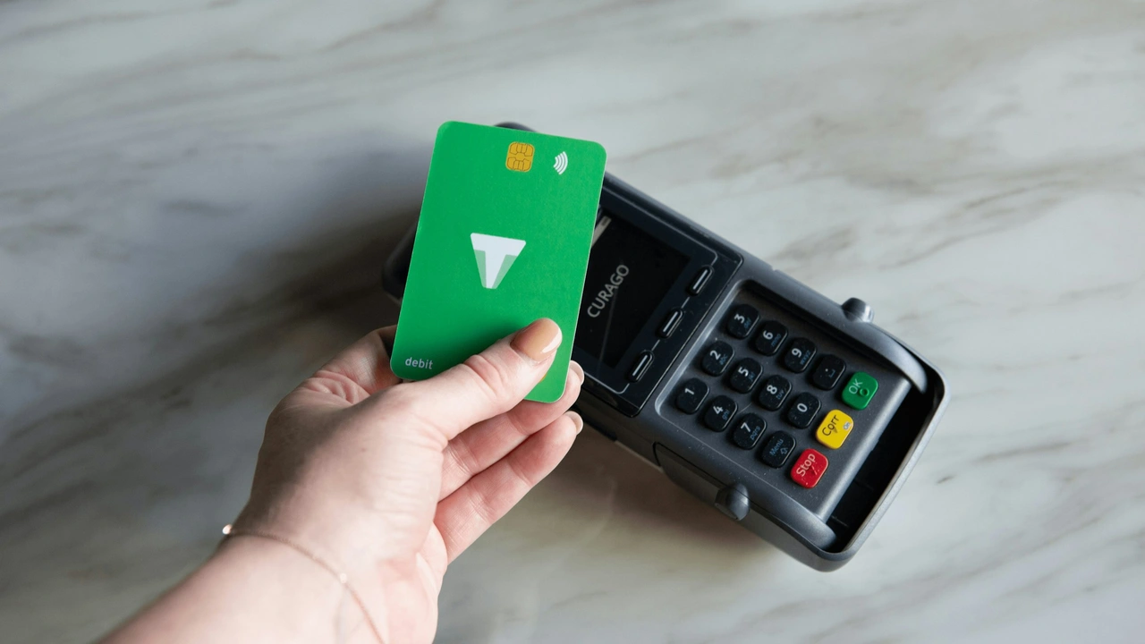 Hand holding a green contactless debit card making a tap-to-pay transaction on a POS card payment terminal at a checkout counter.
