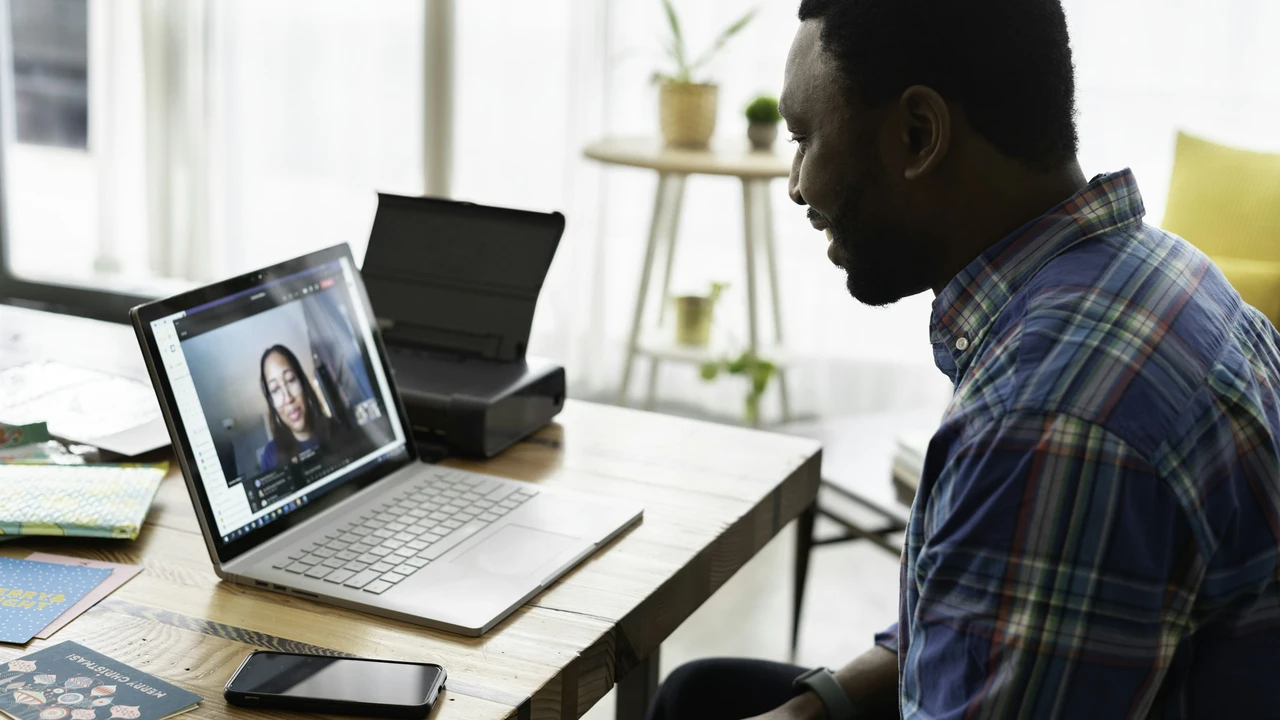 Man attending a video call on a laptop while working from home, sitting at a desk with smartphone and notebooks in a modern home office.