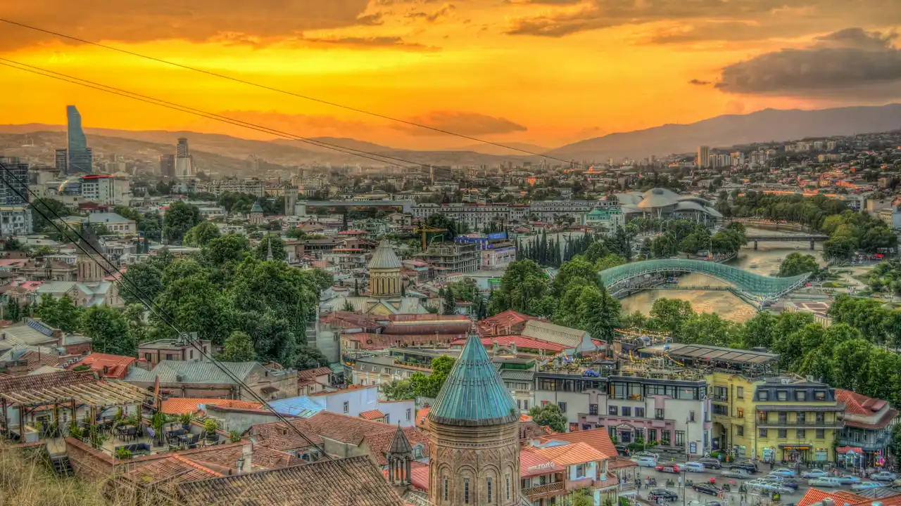 Panoramic view of Tbilisi cityscape at sunset with Kura River, Peace Bridge, historic Old Town rooftops and mountains in the background