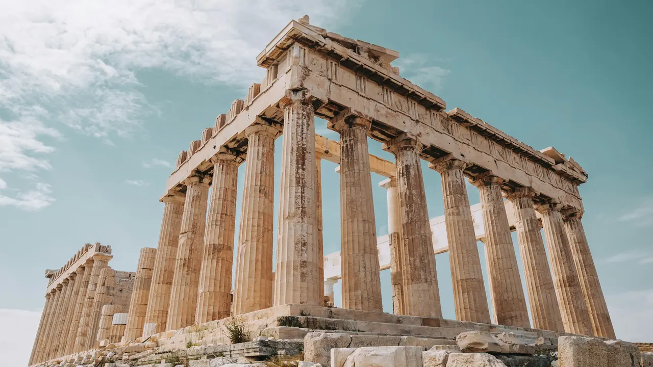 A wide-angle, panoramic photograph of the Parthenon, an ancient Greek temple dedicated to the goddess Athena, located on the rocky outcrop of the Acropolis in Athens. The image captures the iconic Doric-style marble columns and the weathered ruins of the temple's structure under a clear sky, illustrating one of the most famous landmarks of classical Greek architecture.
