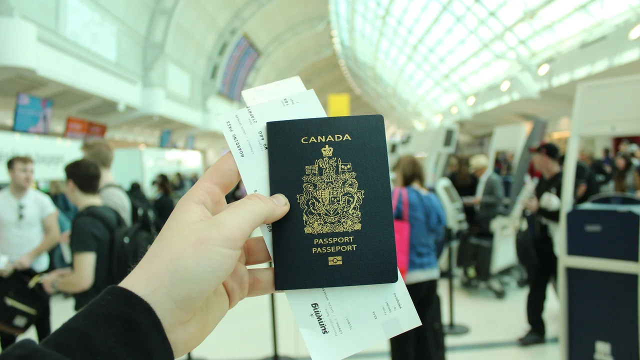 Hand holding a Canadian passport and airline boarding passes inside a busy airport terminal, with travelers and check-in counters in the background.