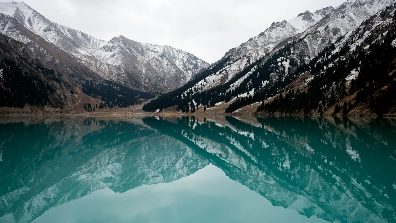 A scenic landscape of the Big Almaty Lake in Kazakhstan, showing turquoise water perfectly reflecting the surrounding snow-capped mountains and evergreen forests under an overcast sky