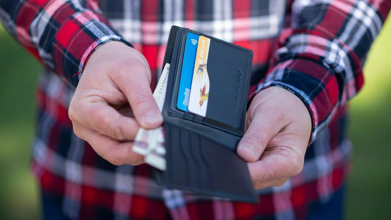 A close-up of a person in a red-and-black plaid shirt holding an open black leather wallet, showing several credit cards and paper banknotes tucked inside.