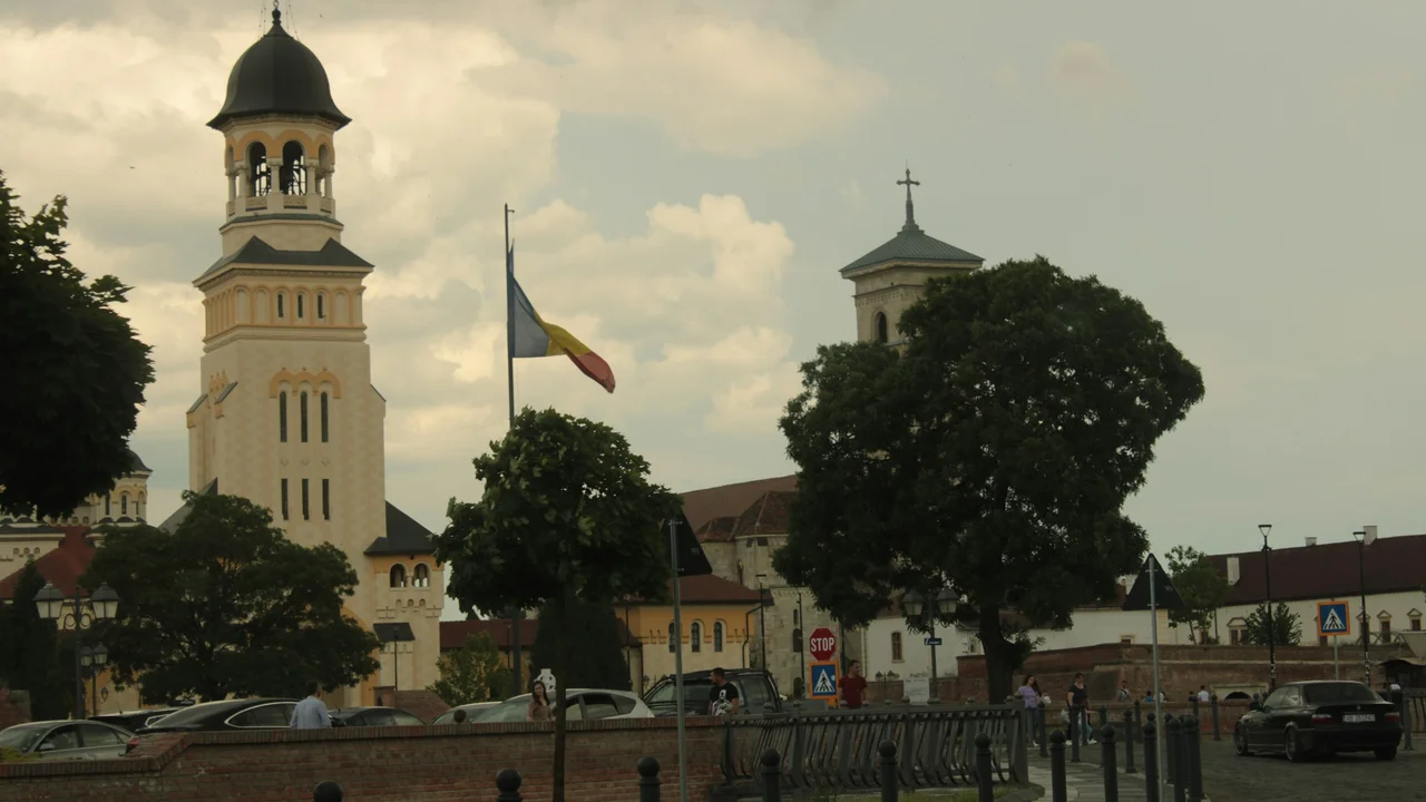 A scenic view of a city square in Romania, featuring a prominent white cathedral with a dark domed bell tower. A Romanian flag flies on a tall flagpole in the center of the square, surrounded by green trees, parked cars, and historic buildings under a cloudy sky