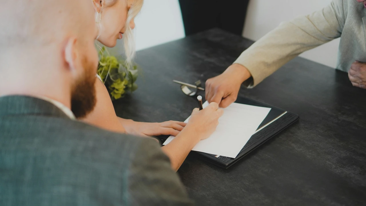 A close-up, high-angle shot of a person signing a white document on a dark desk during a professional meeting. Two other individuals are visible: a man with a beard in the foreground and a person in a tan blazer pointing to the signature line.