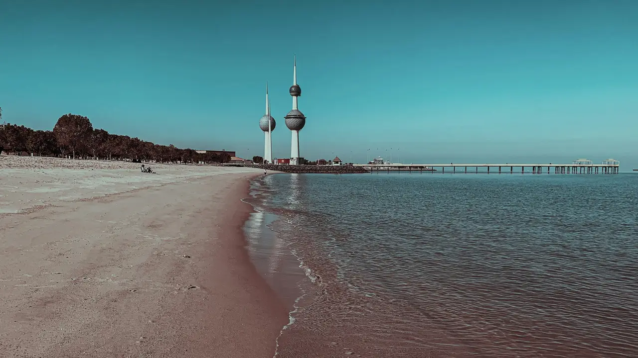 An aerial view of the Kuwait City skyline during sunset, featuring the iconic Kuwait Towers prominently in the foreground. The three slender towers are situated on a promontory overlooking the Arabian Gulf, with their spheres reflecting a warm orange and pink glow from the setting sun.