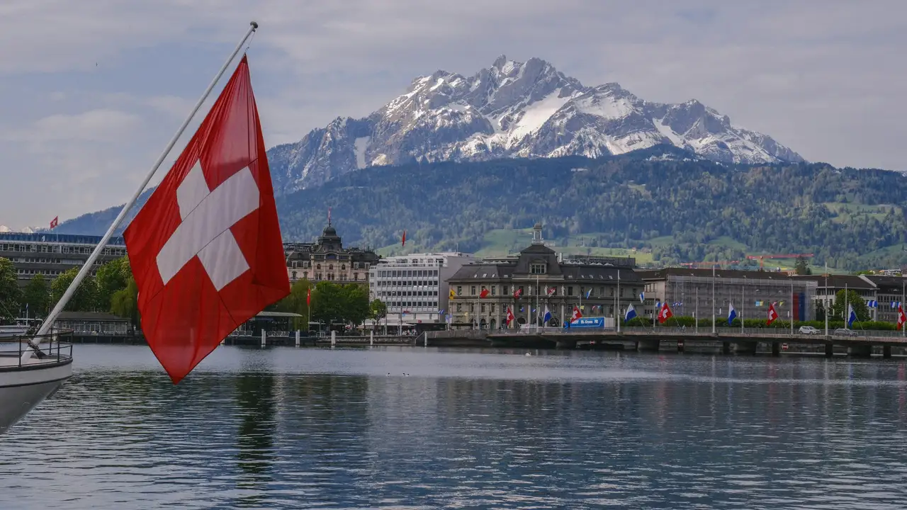 A red Swiss flag with a white cross flutters in the foreground on a boat, overlooking a calm lake and the city of Lucerne, Switzerland, with the snow-capped peaks of Mount Pilatus rising majestically in the background.