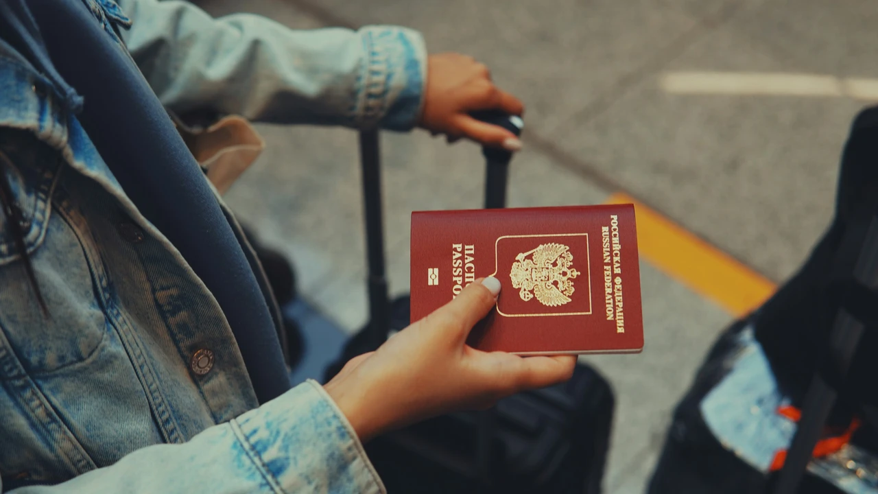 A person wearing a denim jacket holds a red Russian Federation passport while resting their other hand on the handle of a black rolling suitcase. The background shows a paved ground surface, suggesting a traveler at an airport or station.