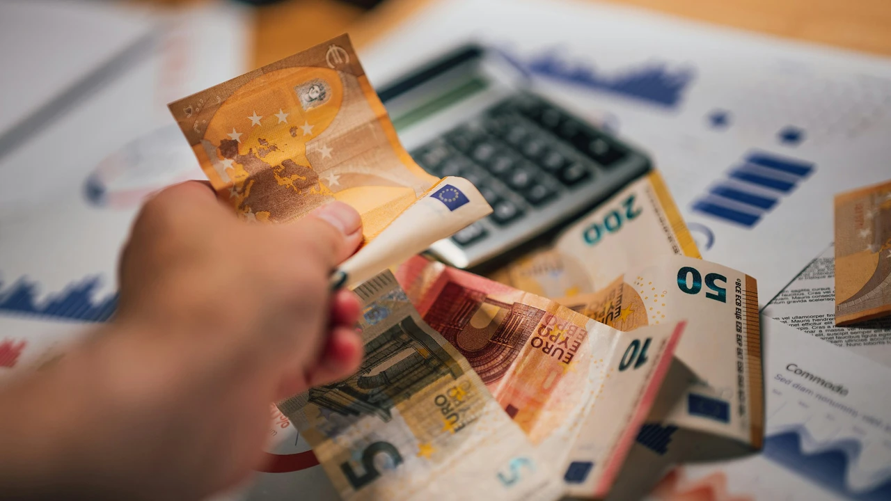 A close-up, slightly blurred shot of a hand holding several Euro banknotes over a desk. In the background, a calculator and financial documents with blue graphs and charts are visible, suggesting a theme of fees or financial processing.