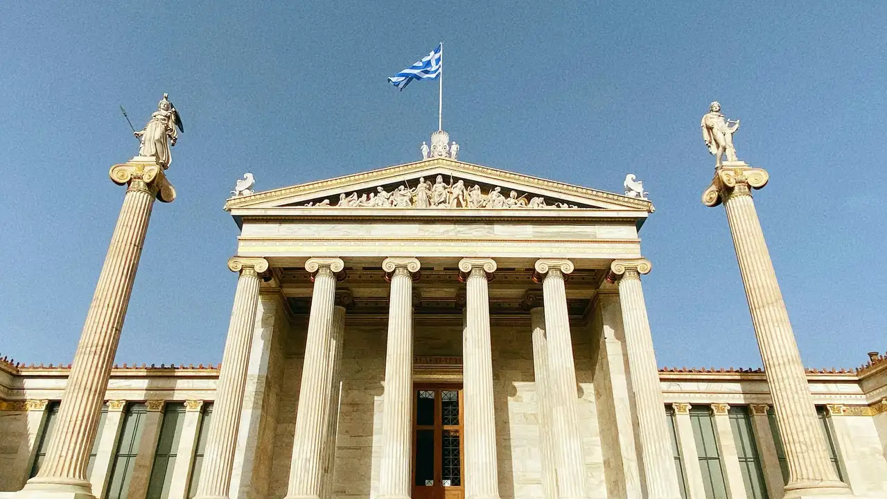 A low-angle shot of the Academy of Athens, a Neoclassical building in Greece. The facade features a row of tall Ionic columns and a pediment filled with sculptures. On either side, high pillars support statues of Athena and Apollo. The Greek flag flies prominently from the center of the roof against a clear blue sky.